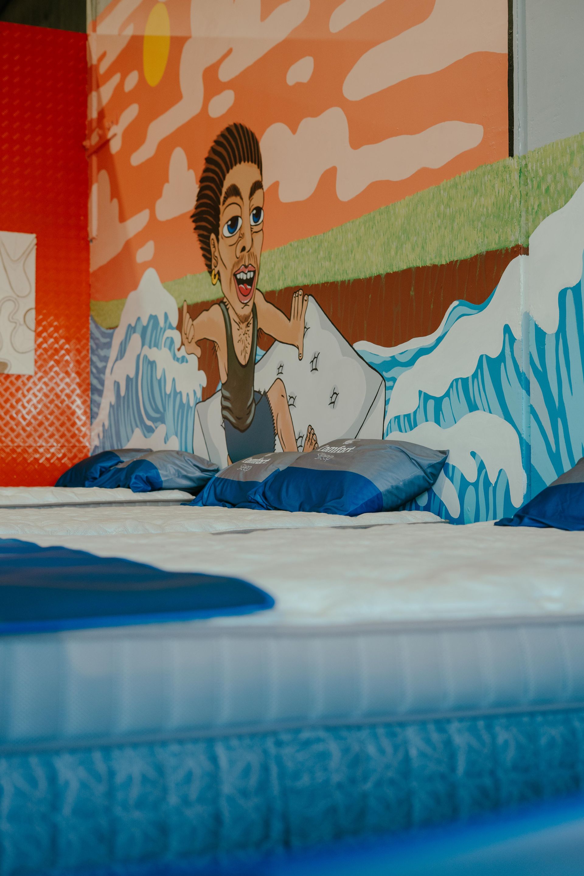 Cartoon figure standing in front of waves painted on a wall, with pillows on a mattress — Coastal Mattress' Direct in Brisbane, QLD  