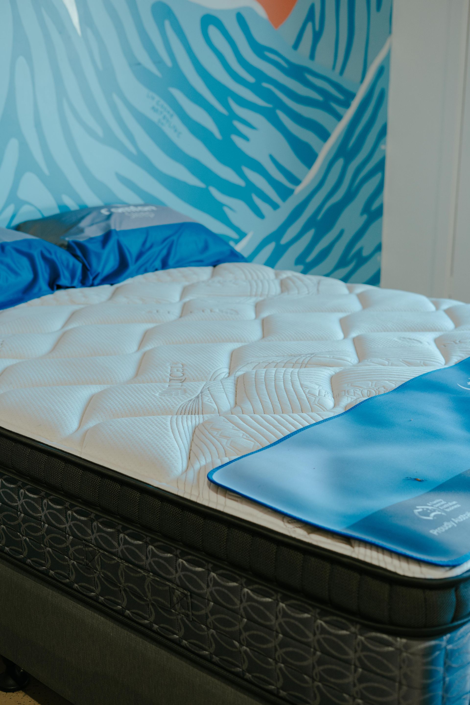 Bed with a white quilted mattress, blue pillows, and a blue decorative sheet, with a blue mural in the background — Coastal Mattress' Direct in South Nowra, NSW