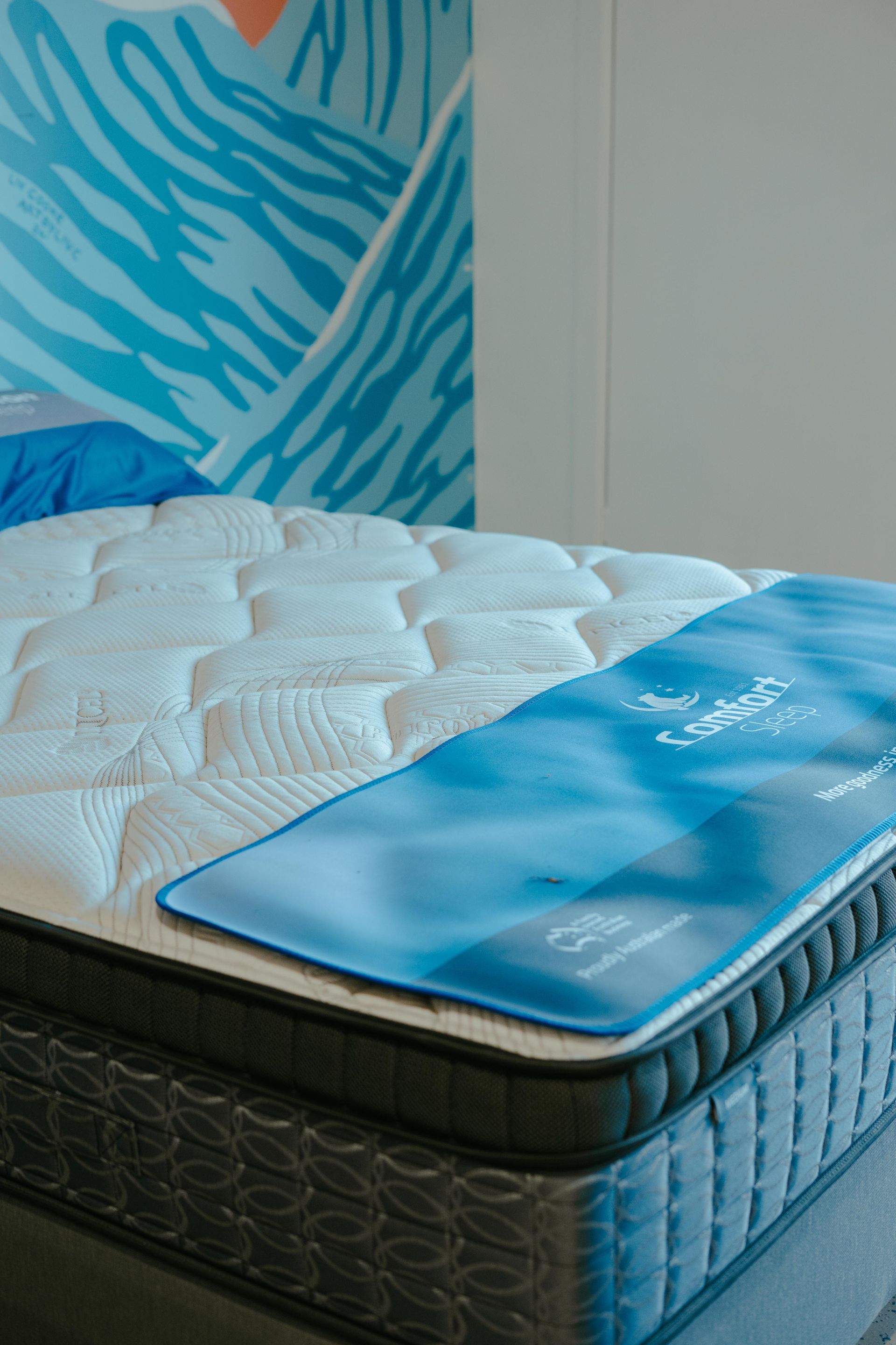 Mattress with white quilted top, blue pillow and promotional overlay; against blue wall art — Coastal Mattress' Direct in Brisbane, QLD  