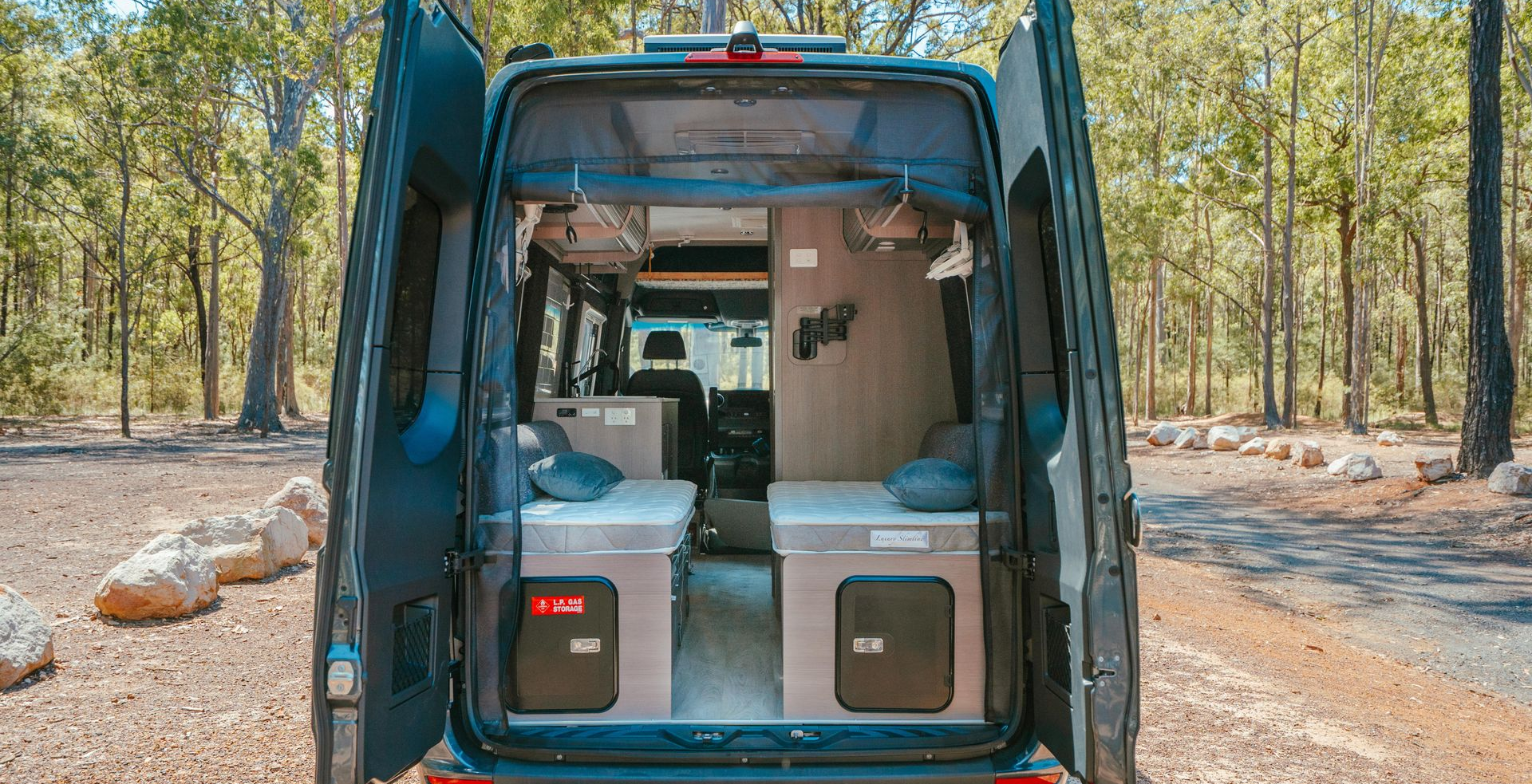 Rear view of a campervan with open doors, revealing a small interior with storage and beds, parked in a wooded area.