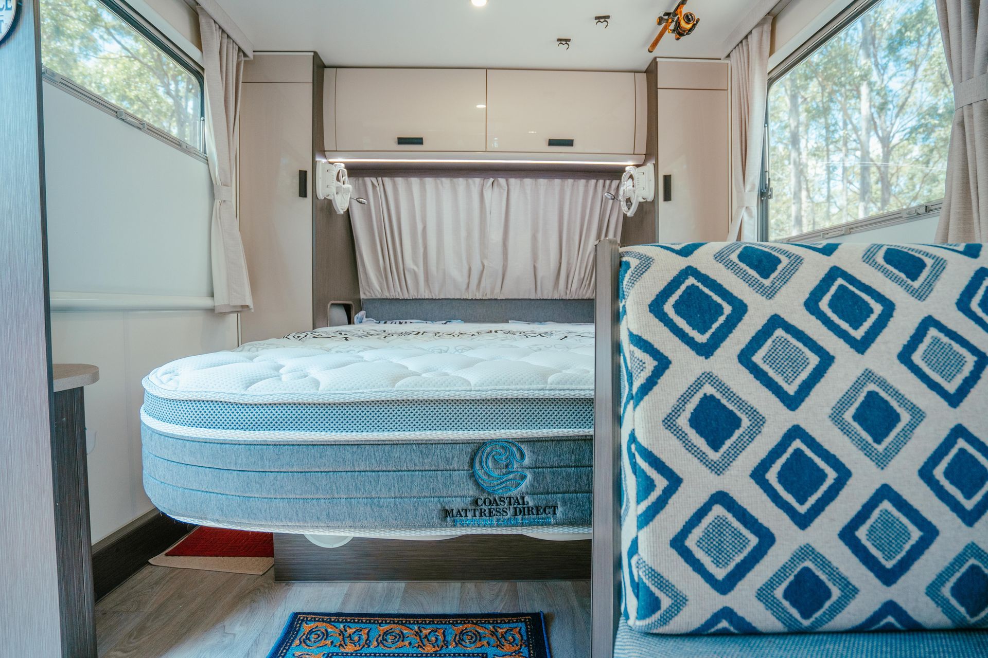 Interior of a camper, showing a bed with a gray mattress, a patterned cushion, and windows.