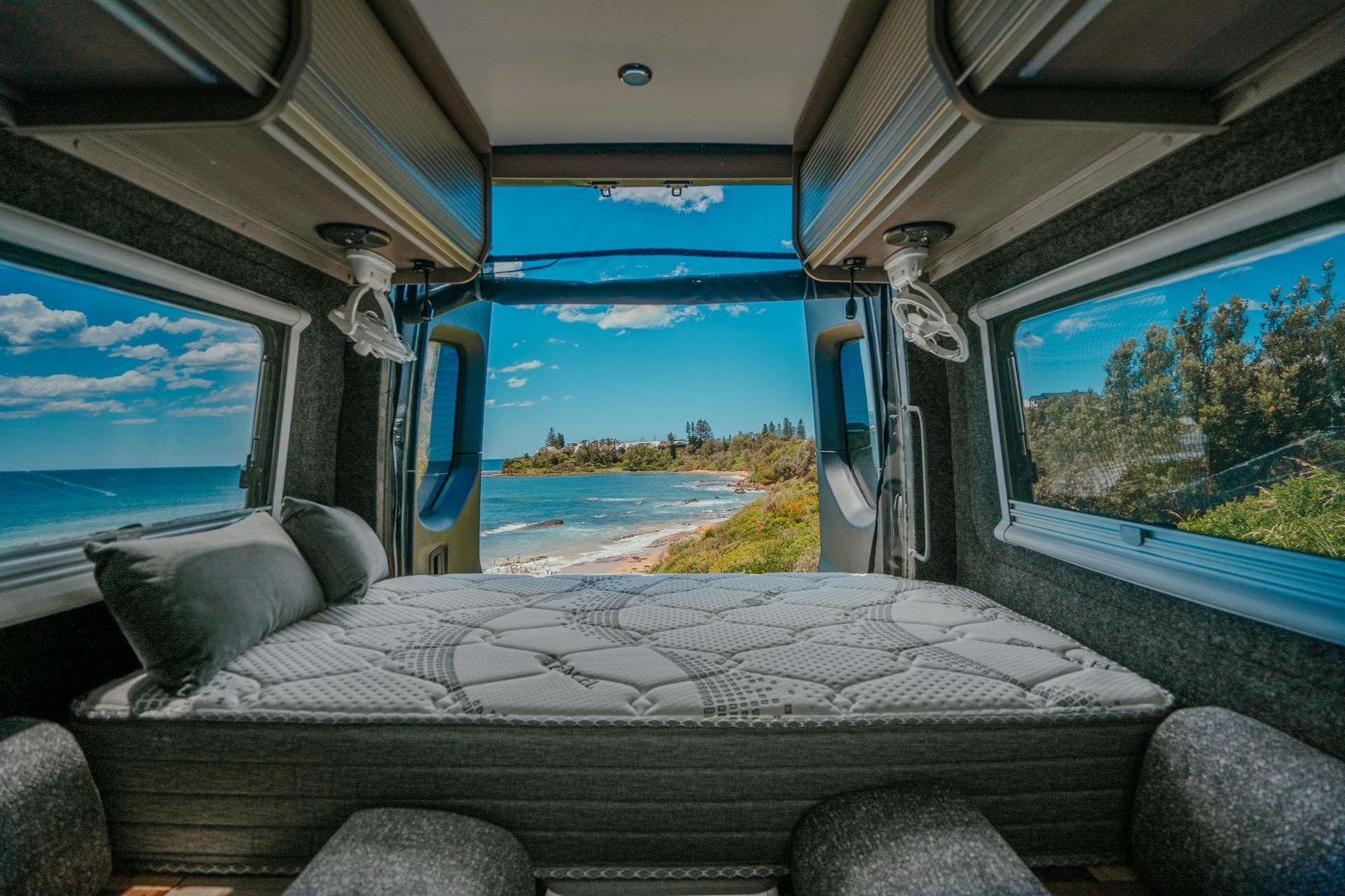Two beds in a camper looking out at a beach, blue sky and water visible.