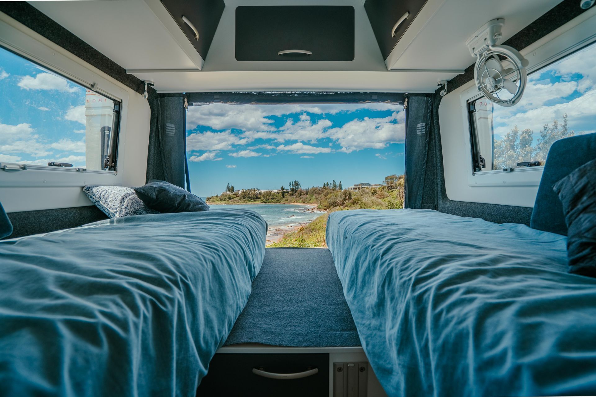 Two beds in a camper looking out at a beach, blue sky and water visible.