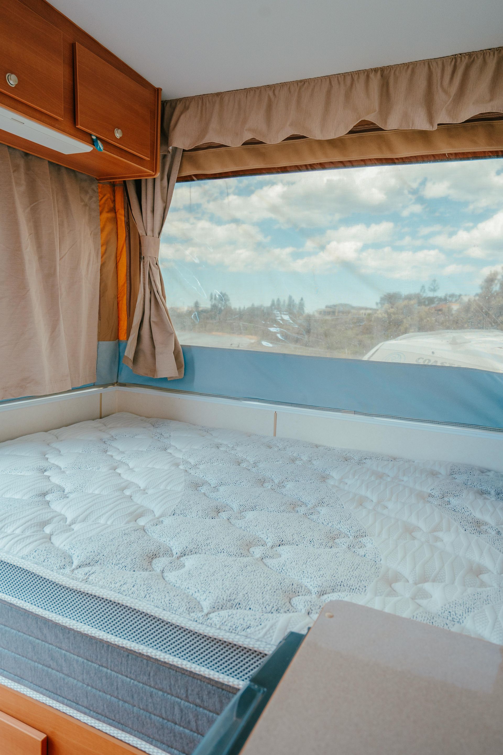 Interior of a pop-up camper with a bed, window, curtains, and wooden cabinets.