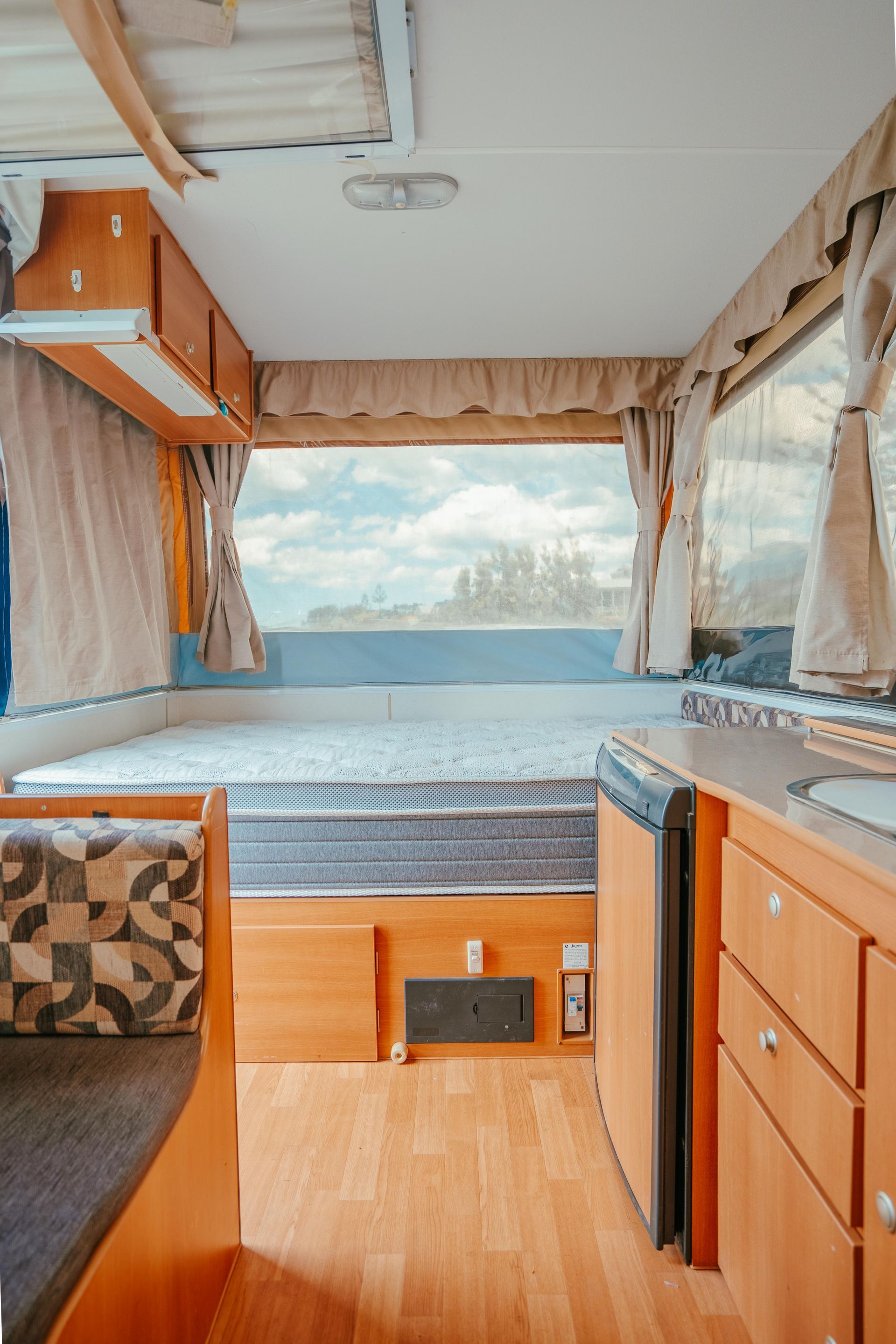 Interior of a camper, showing bed, kitchen area with cabinets and fridge, and a large window.