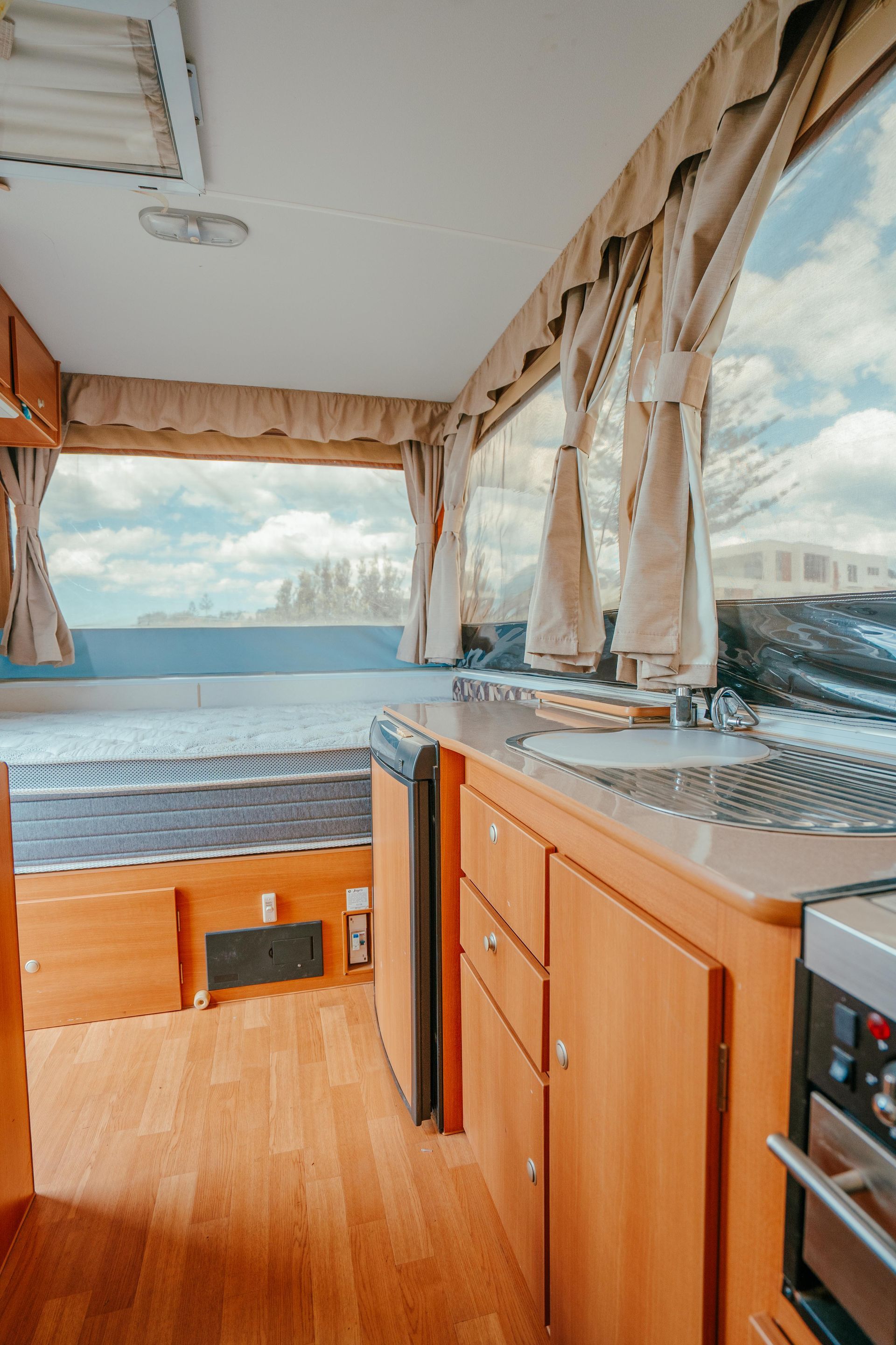 Interior of a camper, featuring kitchen, bed, and window with a view of the outdoors. Wooden cabinets and flooring — Coastal Mattress' Direct in Brisbane, QLD