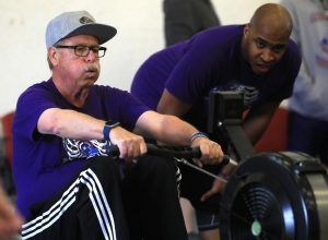 A man is riding a rowing machine while another man watches.
