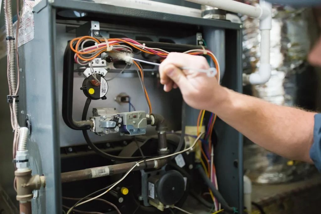 Person working on furnace, using a screwdriver; wires, gas valves visible inside unit.
