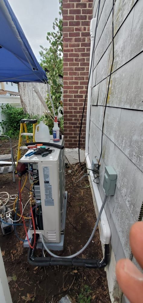HVAC unit being worked on, next to brick chimney and building. Ground, ladder, tools, and blue tarp are visible.