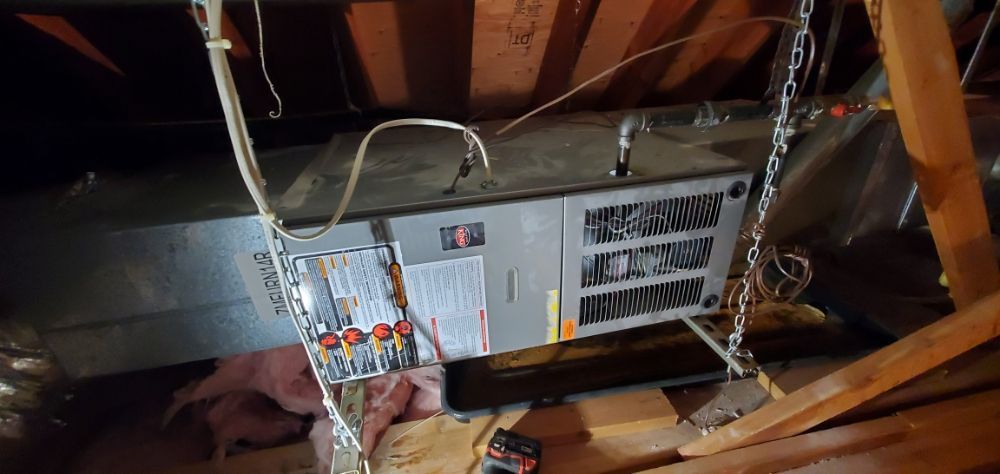 HVAC unit suspended in an attic. Wires and chains present. Wooden beams and insulation visible.