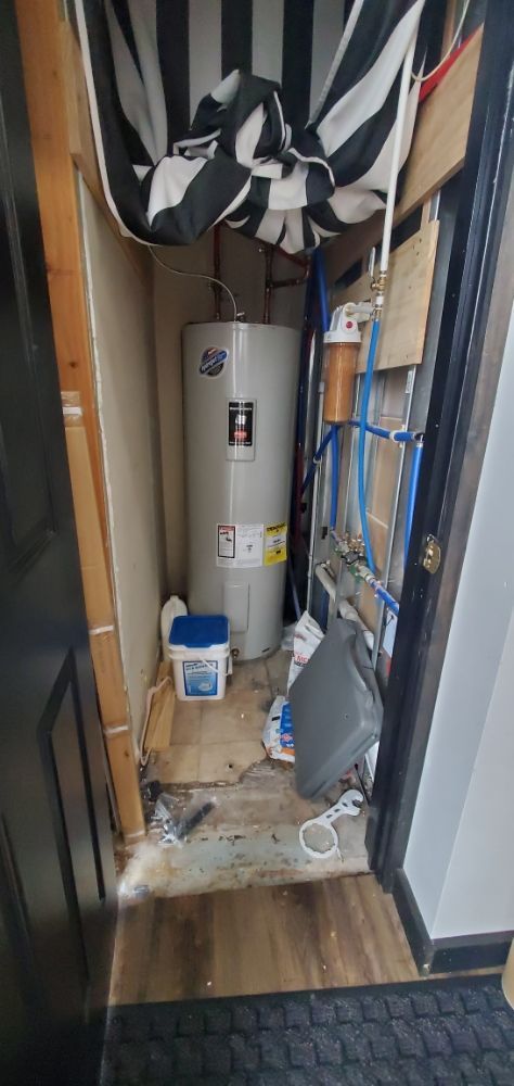 A water heater sits inside a small storage room, with pipes and other items visible.