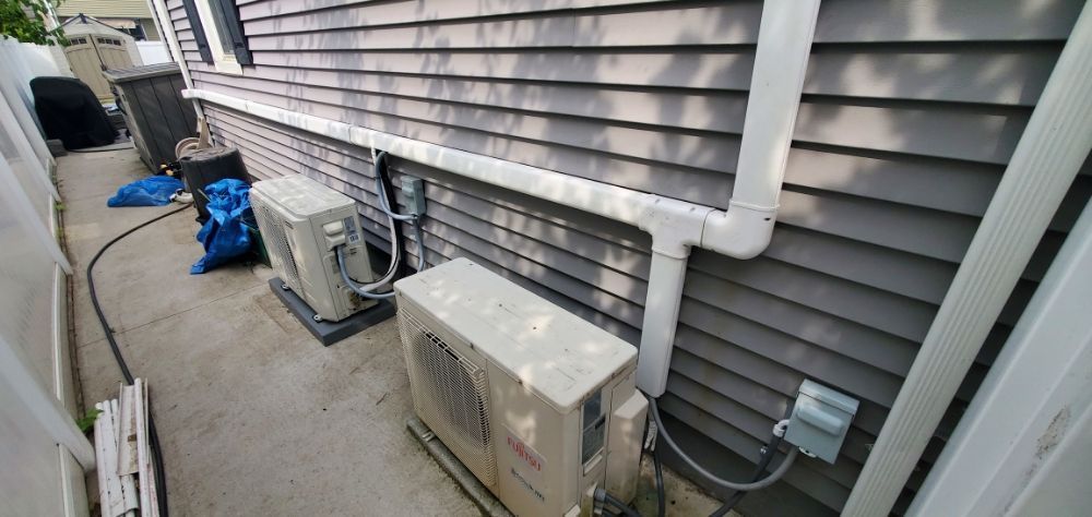 Air conditioning units along a gray siding house. A tight walkway is between the house and a fence.