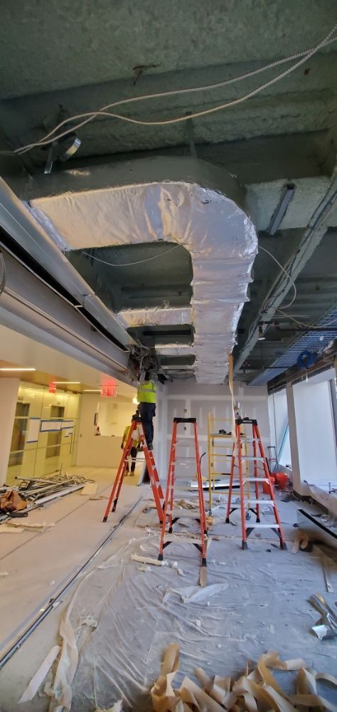 Construction site with workers on ladders; ductwork wrapped in white, unfinished interior.