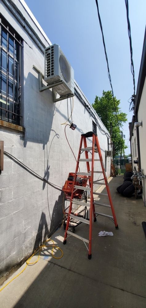 An air conditioning unit on a white brick building with a ladder beside it under a blue sky.