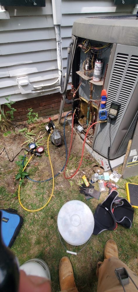 A person working on an outdoor air conditioning unit. Gauges, wires, and tools are visible.