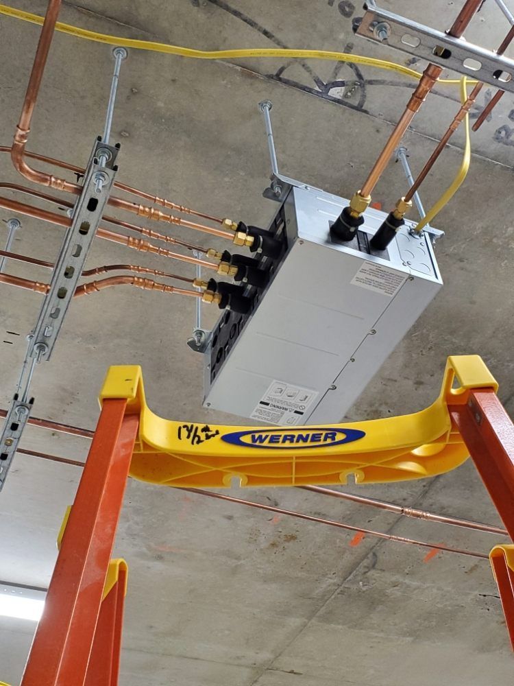 A silver electrical unit is suspended from the ceiling. A yellow ladder supports it. Copper pipes and wiring are visible.