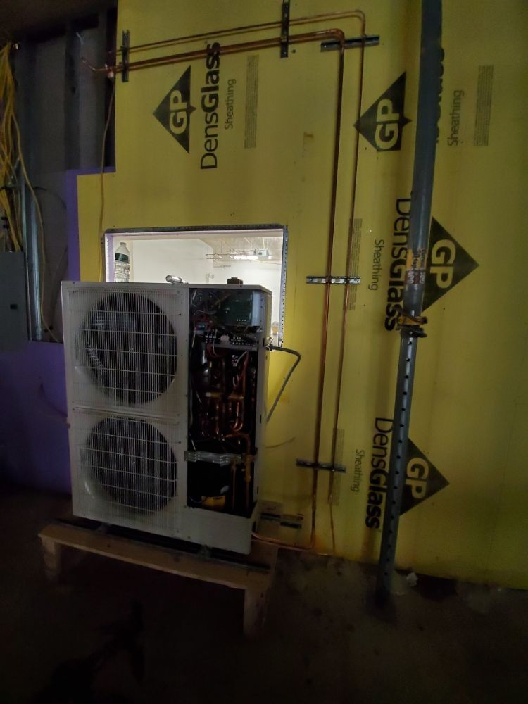 An HVAC unit with copper piping against a yellow wall with exposed insulation.