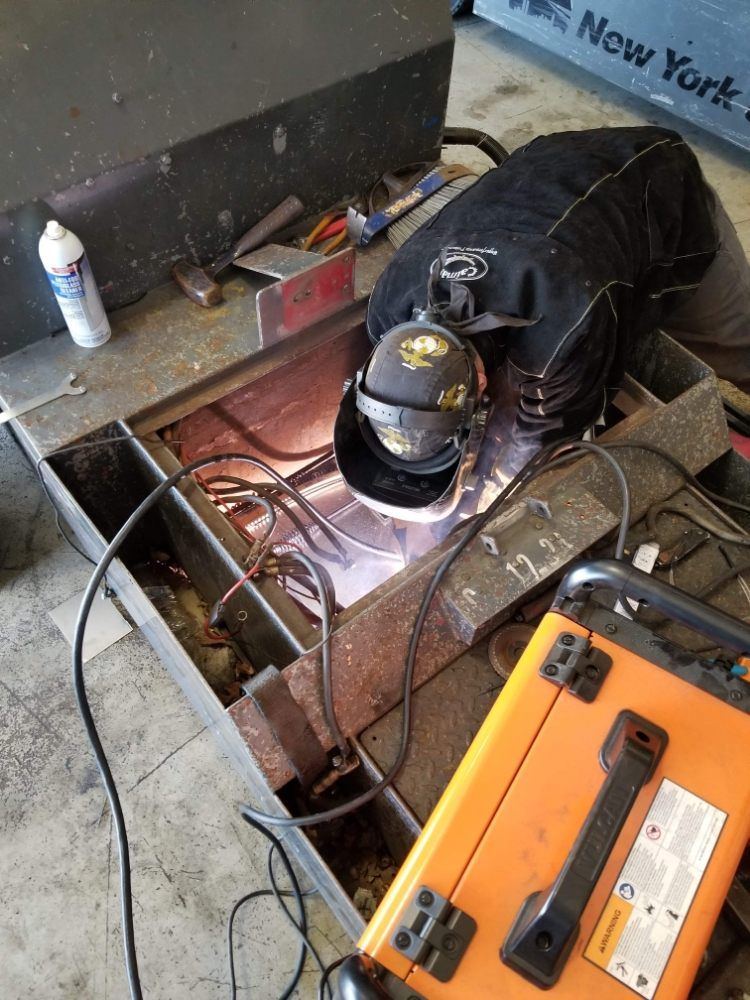 Welder in black protective gear working in a metal frame; orange welding machine in the foreground.