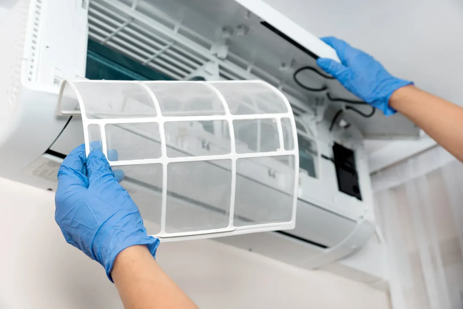 Hands in blue gloves removing an air conditioner filter for cleaning. White unit mounted on a wall.
