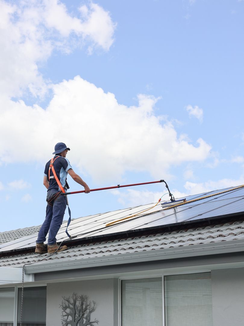 Person cleaning solar panels on a rooftop with safety harness and extension pole — Col Groves Electrics Pty Ltd in Ballina, NSW
