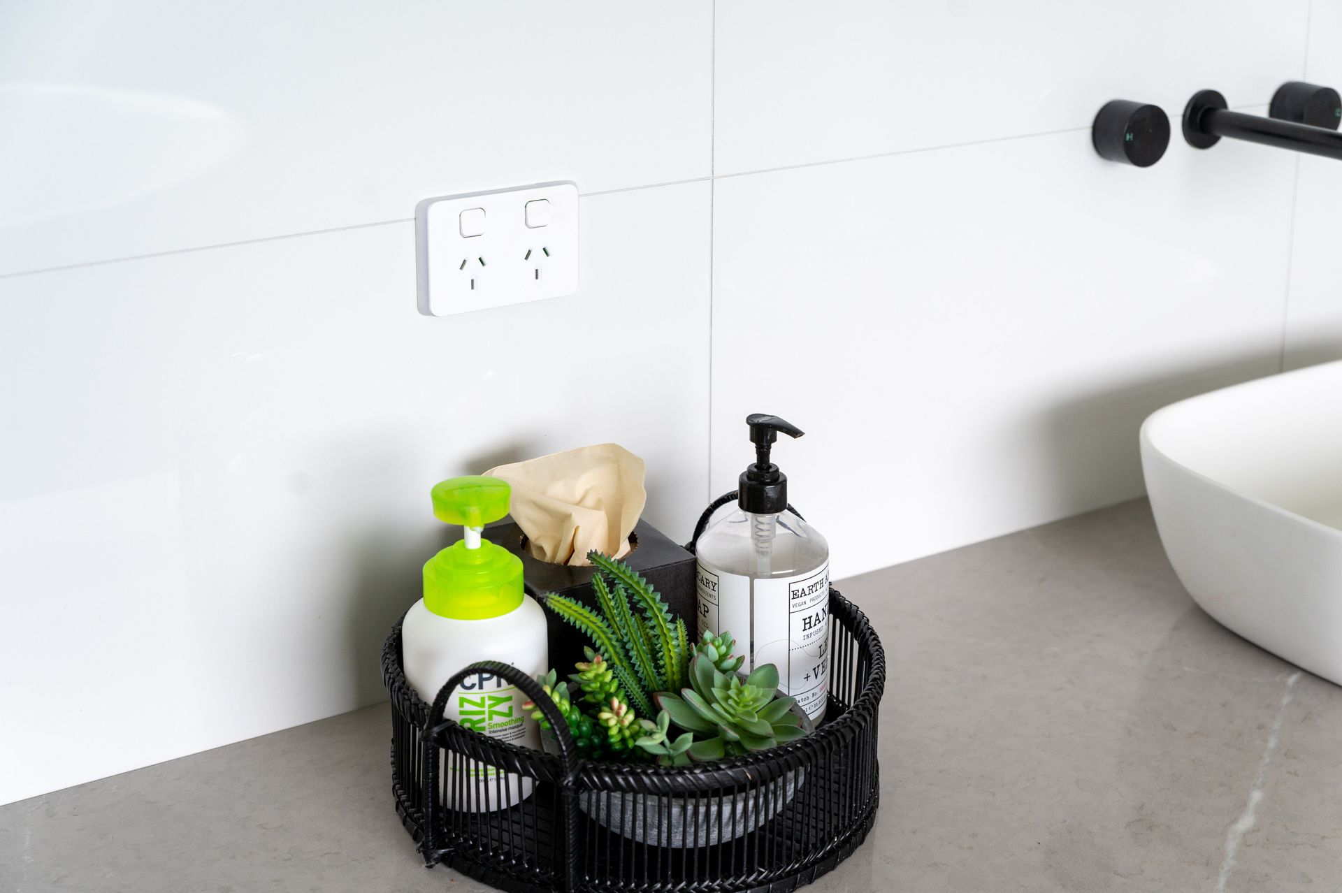 A bathroom counter with a black basket holding soap dispensers, plants, and a power outlet.