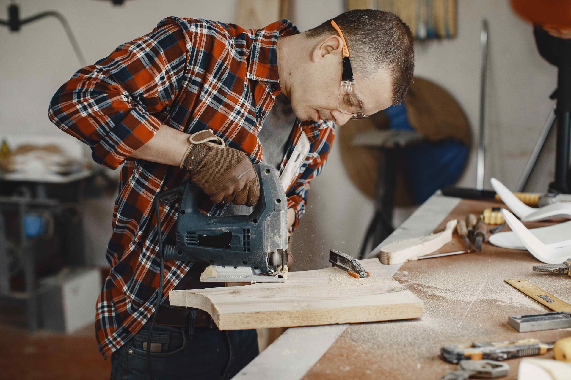 A person in a red plaid shirt and safety glasses uses a jigsaw to cut a piece of wood in a workshop.