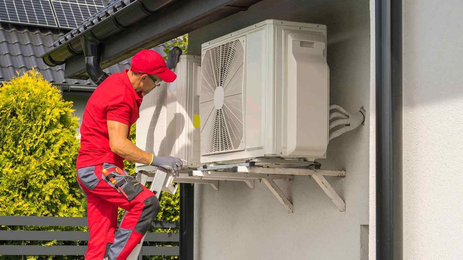 Technician in a red uniform installs or repairs an outdoor air conditioning unit mounted on a building wall.