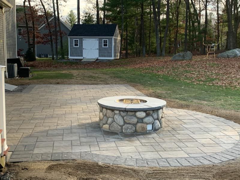 New paver patio with a custom stone fire pit surrounded by a stone wall, in a yard with a shed in the background.