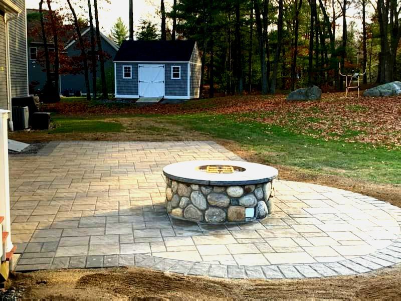 Stone fire pit on a paved patio, with a shed and trees in the background.