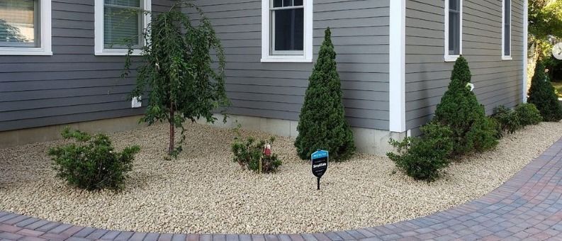Landscaped yard with gravel, shrubs, and trees along a gray house with white trim and windows. Hardscaping project