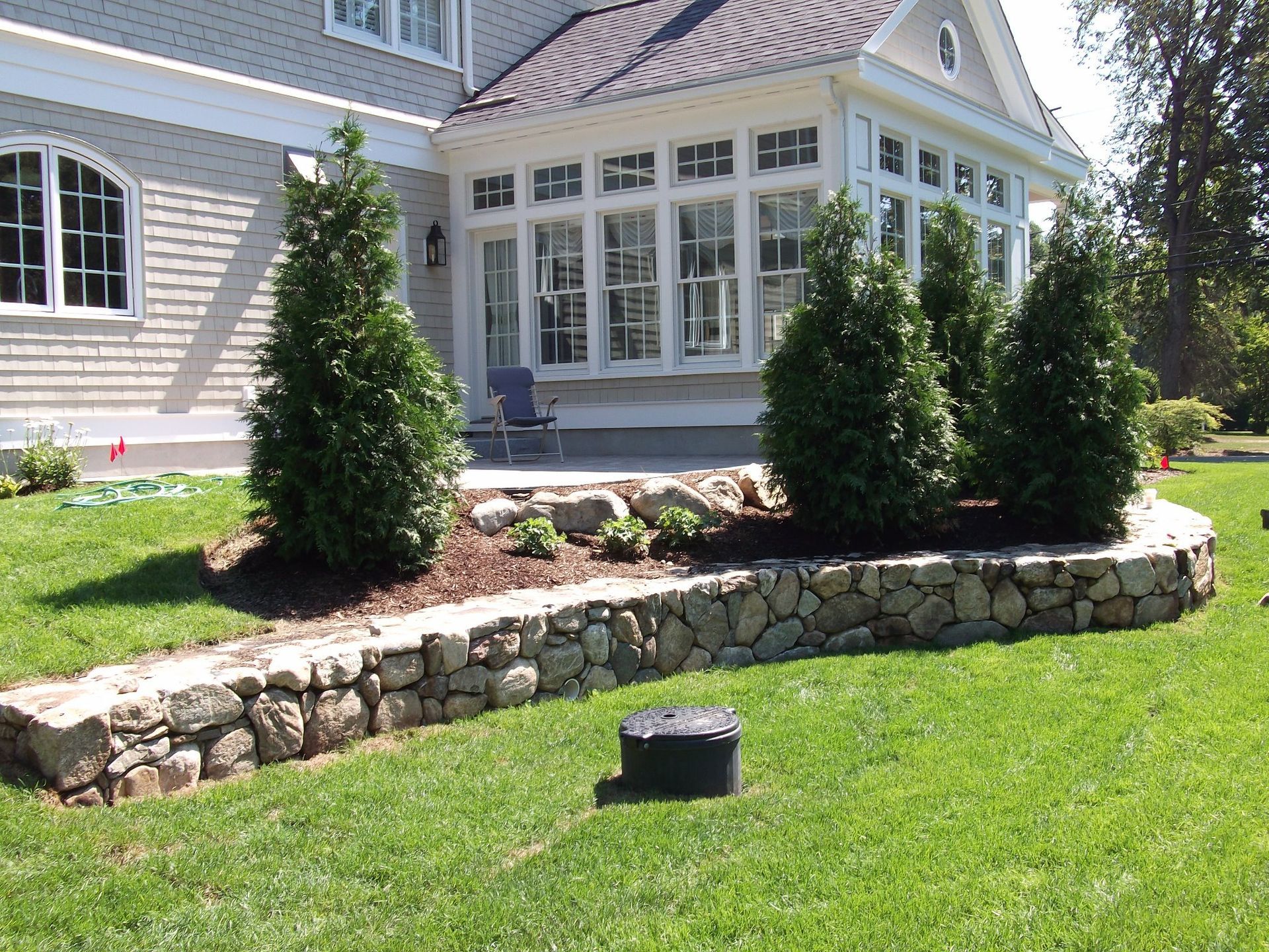 Stone retaining wall with new trees in front of a house. Green lawn and sunny setting.