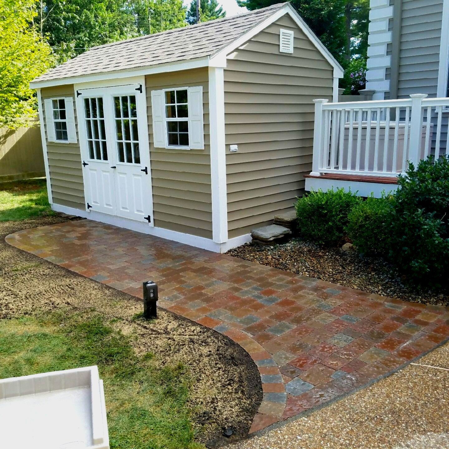 Tan shed with paver pathway and small porch next to a white house.