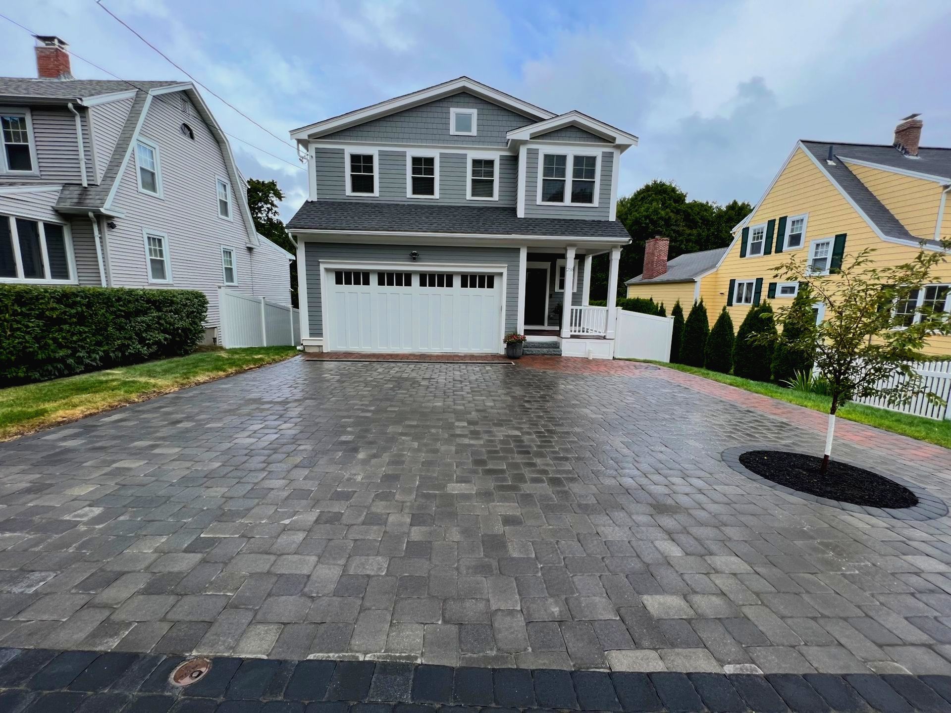 Two-story gray house with white garage door and brick-paved driveway. Paver driveway with brick walkway installed.