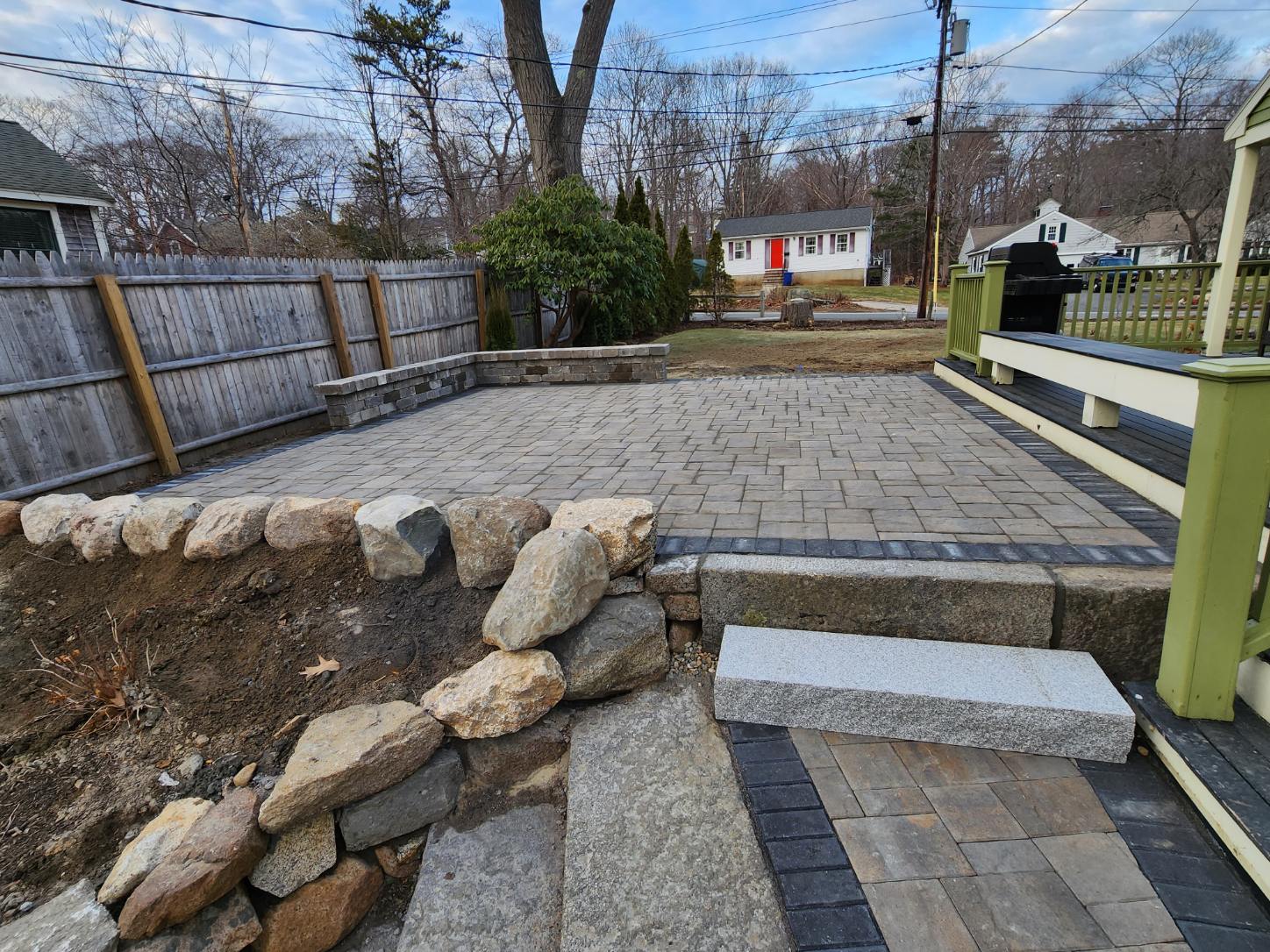 Paver patio with a darker border, rock retaining wall, and wooden fence. Granite step and retaining block wall. 