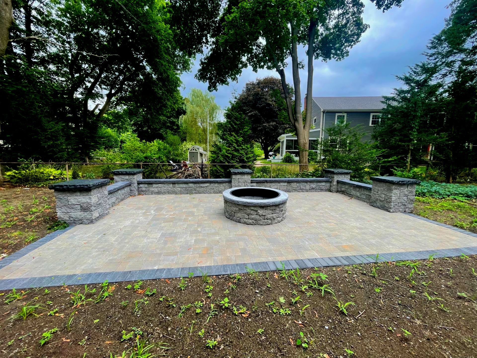 A backyard patio with a fire pit, surrounded by stone walls, trees, and a house in the background. New project. 