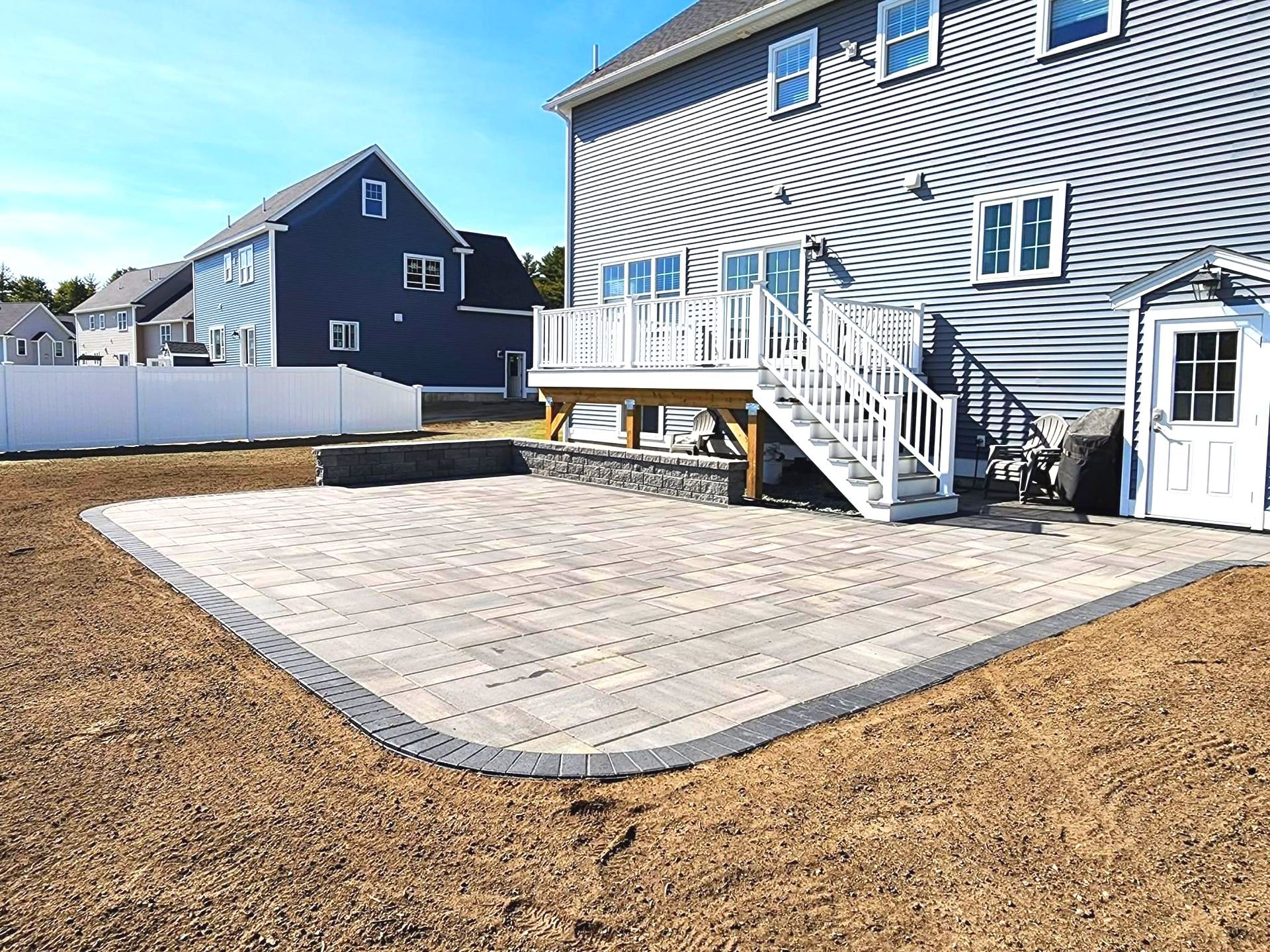 Backyard patio with a house in the background. Pavers, retaining wall, and deck visible.