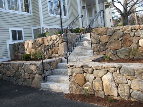 Stone retaining walls with granite steps leading up to a house entrance; black handrails and asphalt driveway.
