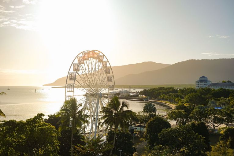 A Ferris wheel stands on Cairns Esplanade at sunrise, with palm trees in the foreground and mountains in the distance.