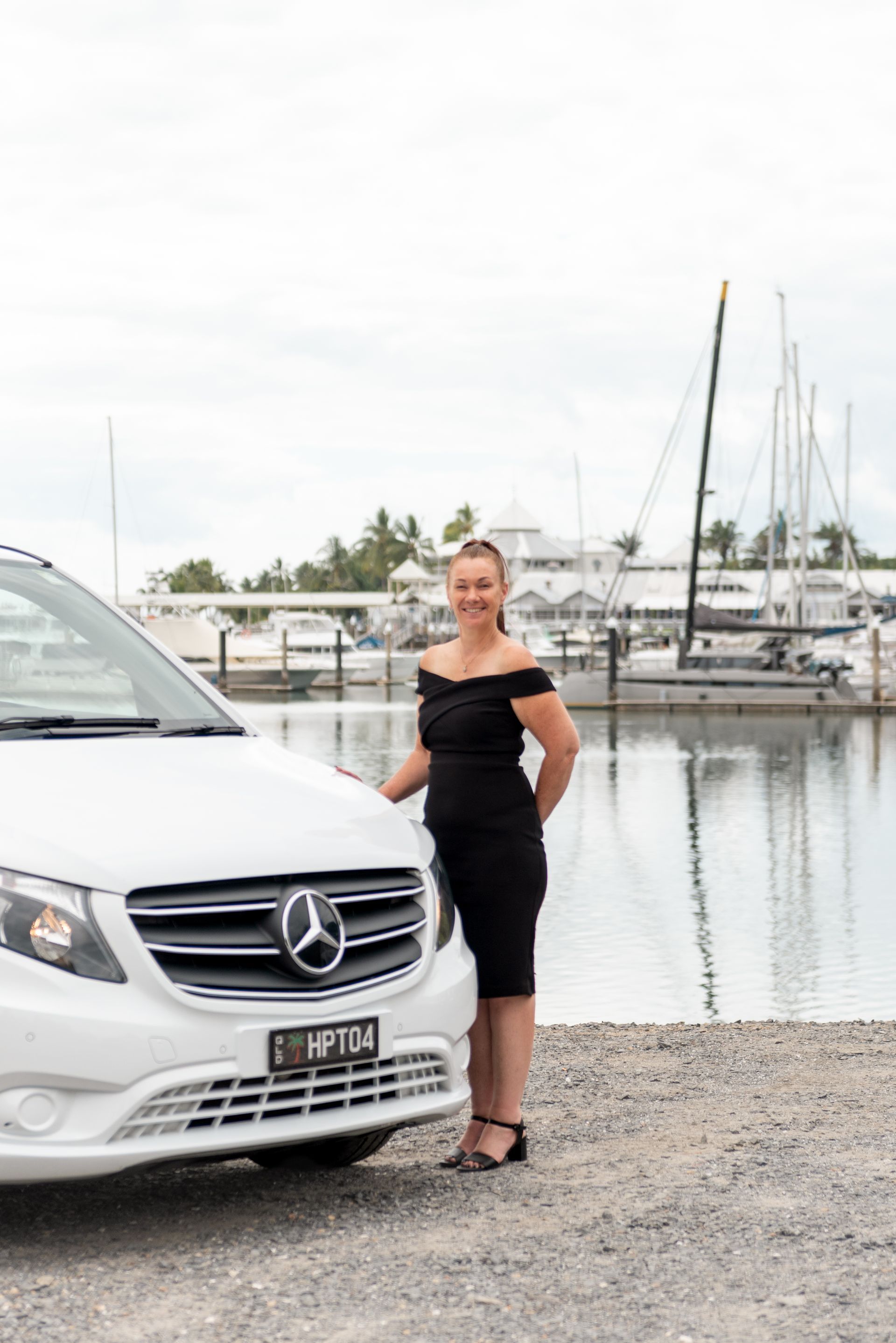 Heather in a black outfit stands beside a white Mercedes van parked at a boat marina.
