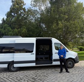 Man in a black suit standing in front of a large black SUV in a city square.