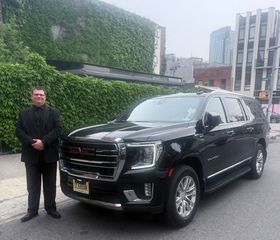Man in a black suit standing in front of a large black SUV in a city square.