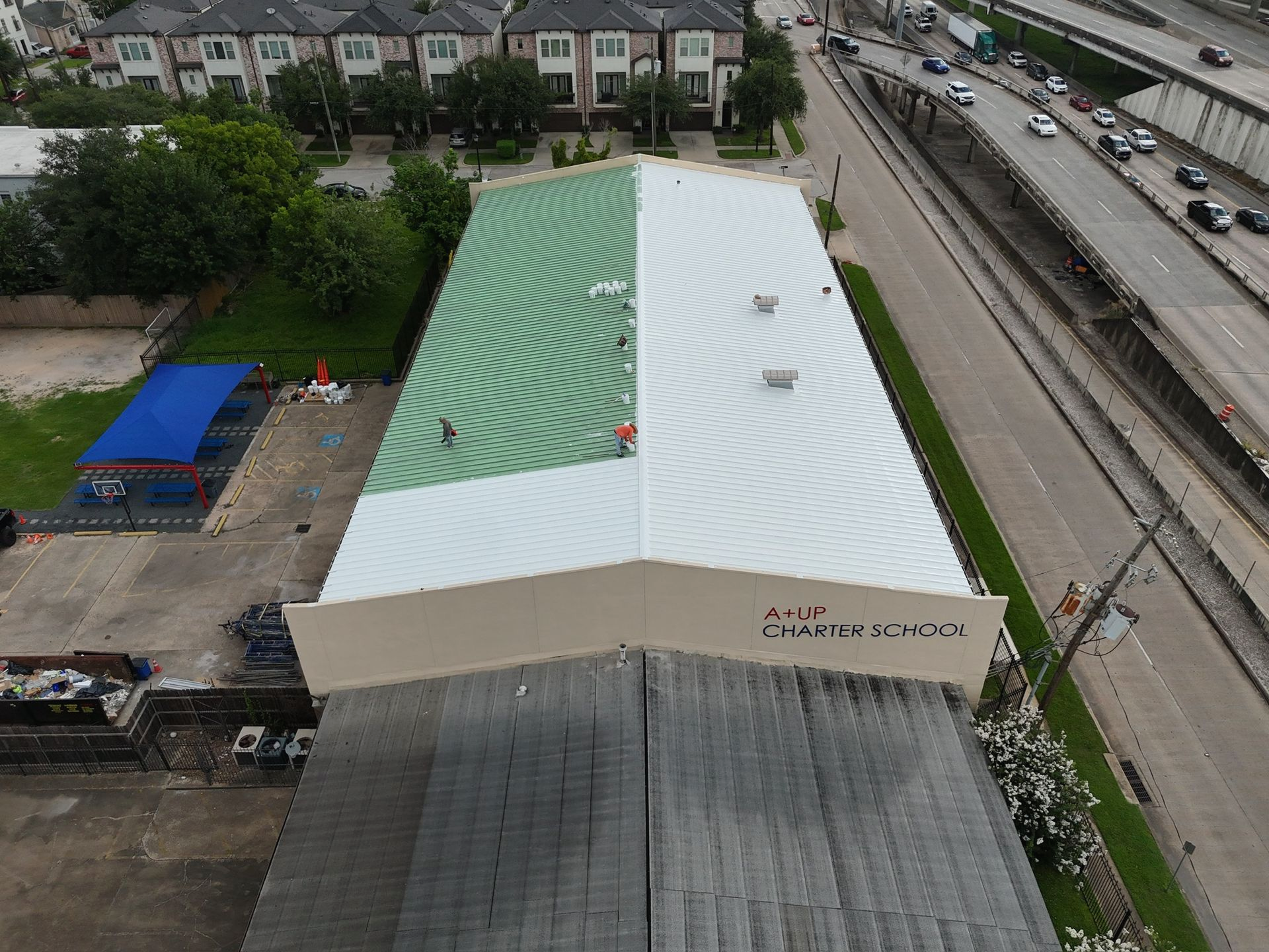 School building with a split roof: half green swirled design, half white. People on roof, highway visible.