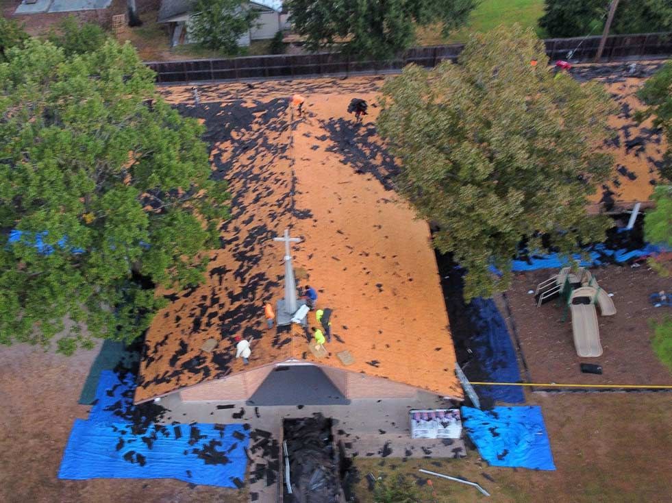 Church roof being repaired after storm damage. Workers on the orange roof, debris, blue tarps, green trees.