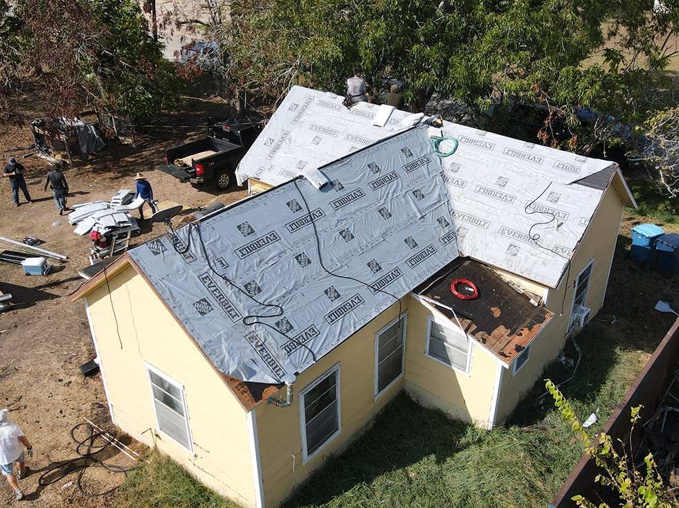 House with partially replaced roof. Roofing workers on the ground and roof. Beige house with brown trim and windows.