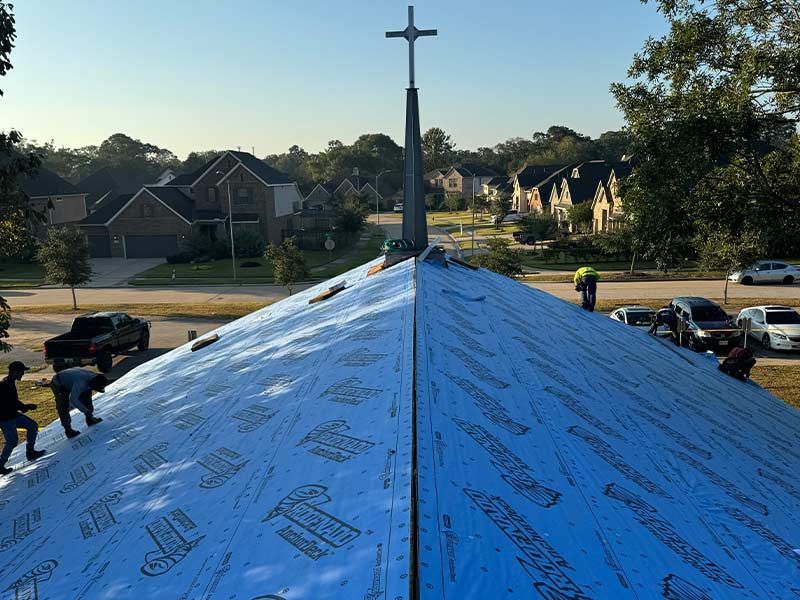 Workers on a church roof installing blue protective underlayment. Houses and cars in the background. A cross on the steeple.