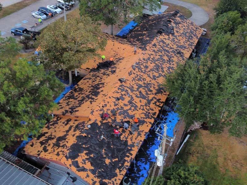 Roof replacement in progress: Workers on a partially stripped roof covered with tarps, trees, and parked cars nearby.