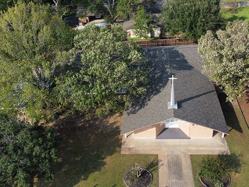 Aerial view of a church with a cross on the roof, surrounded by trees and a driveway.