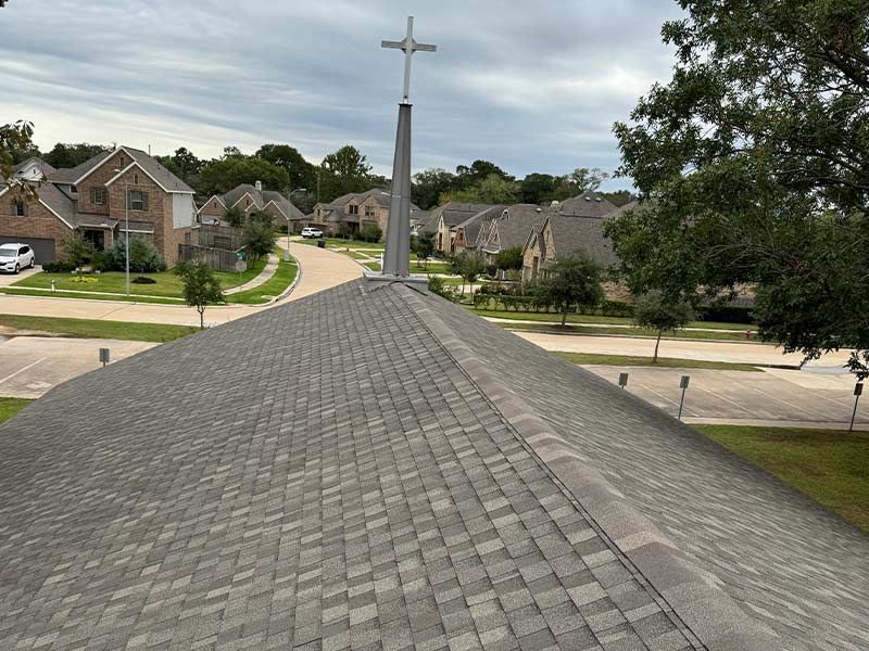 Church roof with cross, overlooking suburban street and houses. Gray shingles and cloudy sky.