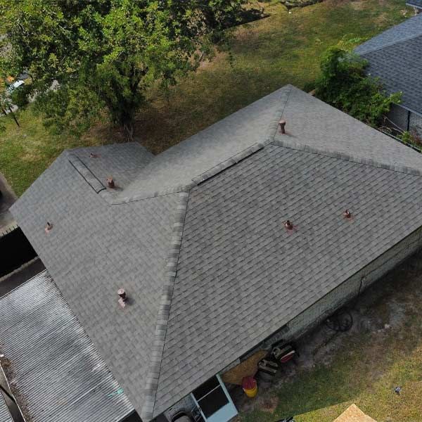 Overhead view of a gray shingle roof on a house, with several vents, set amidst a grassy yard and nearby trees.