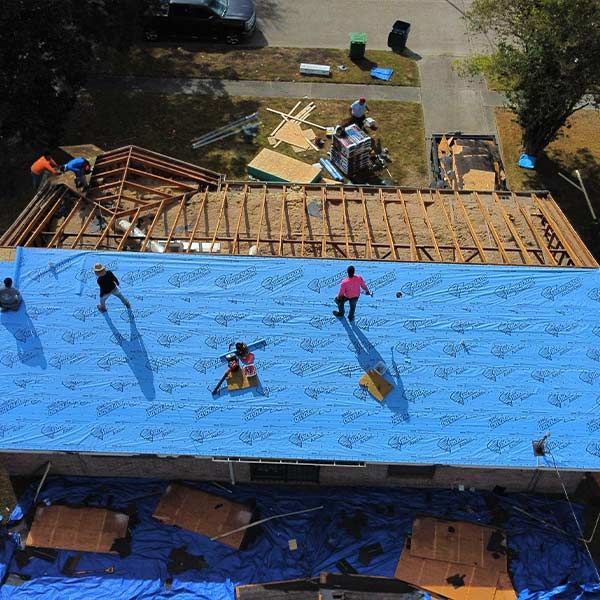 Workers installing blue tarp on a roof. Construction debris and partially exposed roof structure visible.